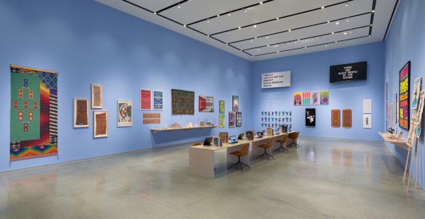 Gallery interior with light blue walls. There are multiple artworks of different mediums and a long wooden desk with books and monitors.