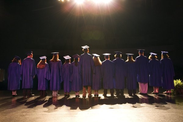 A group of graduates in blue caps and gowns stand on a stage, facing away under bright lights.
