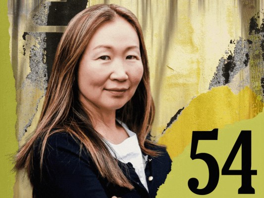 A woman with long hair in a black jacket stands confidently against a yellow and green textured background with the number 54 prominently displayed.