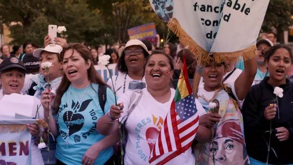 A crowd of people with medium and dark skintones marching and smiling.