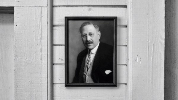 A black and white photograph of a man with a mustache, dressed in a suit and tie, is framed and hanging on a white wooden wall. The man has a serious expression and the setting appears to be formal.