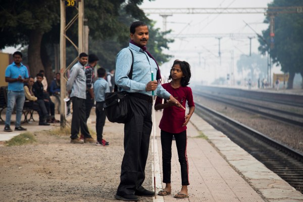 A blind man holding a white cane and a child stand on a train platform. Other people are seated and standing nearby. Train tracks are visible in the background.