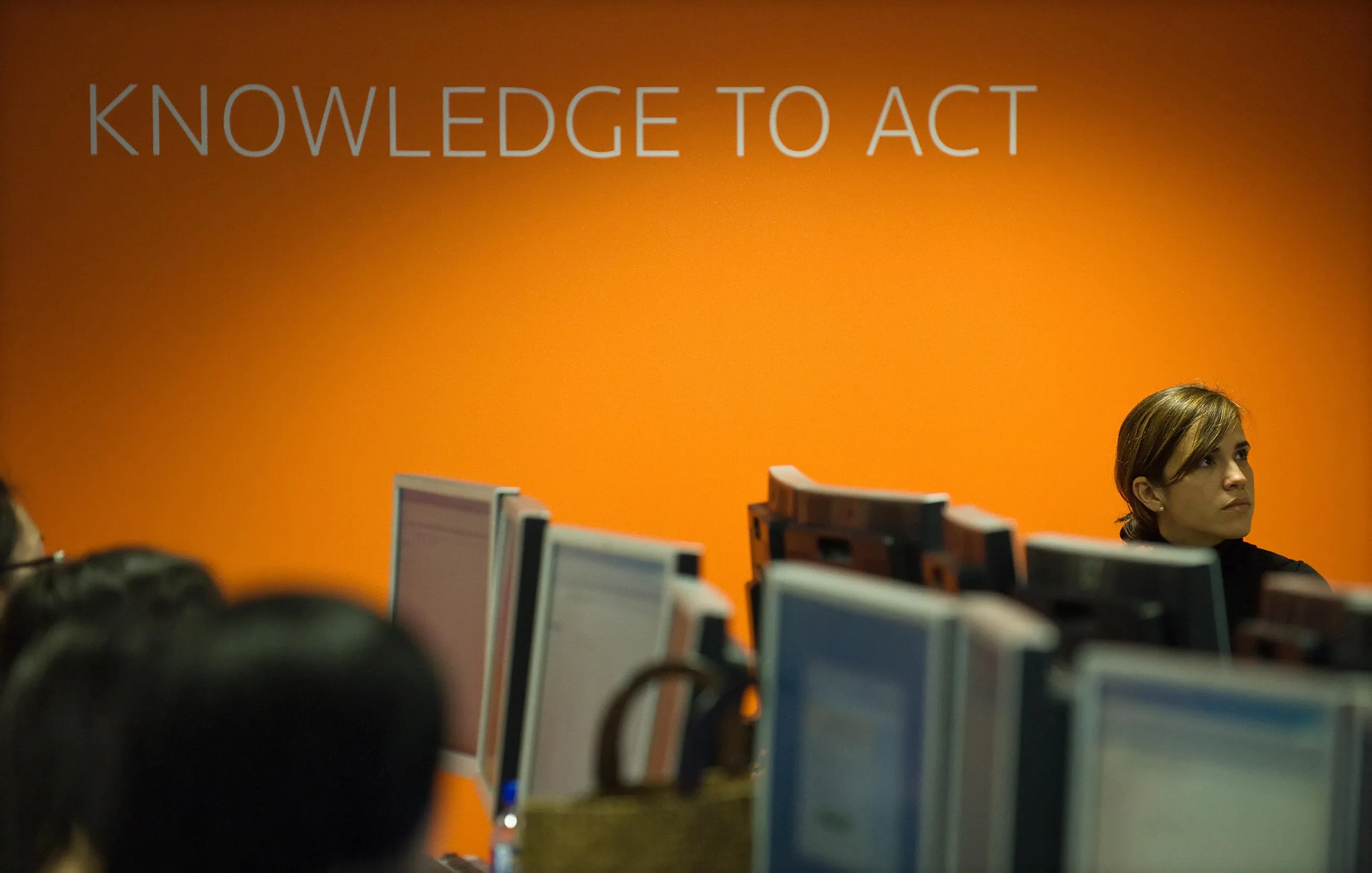 Woman sitting near multiple computer monitors in an office with an orange wall displaying the words "Knowledge to Act.