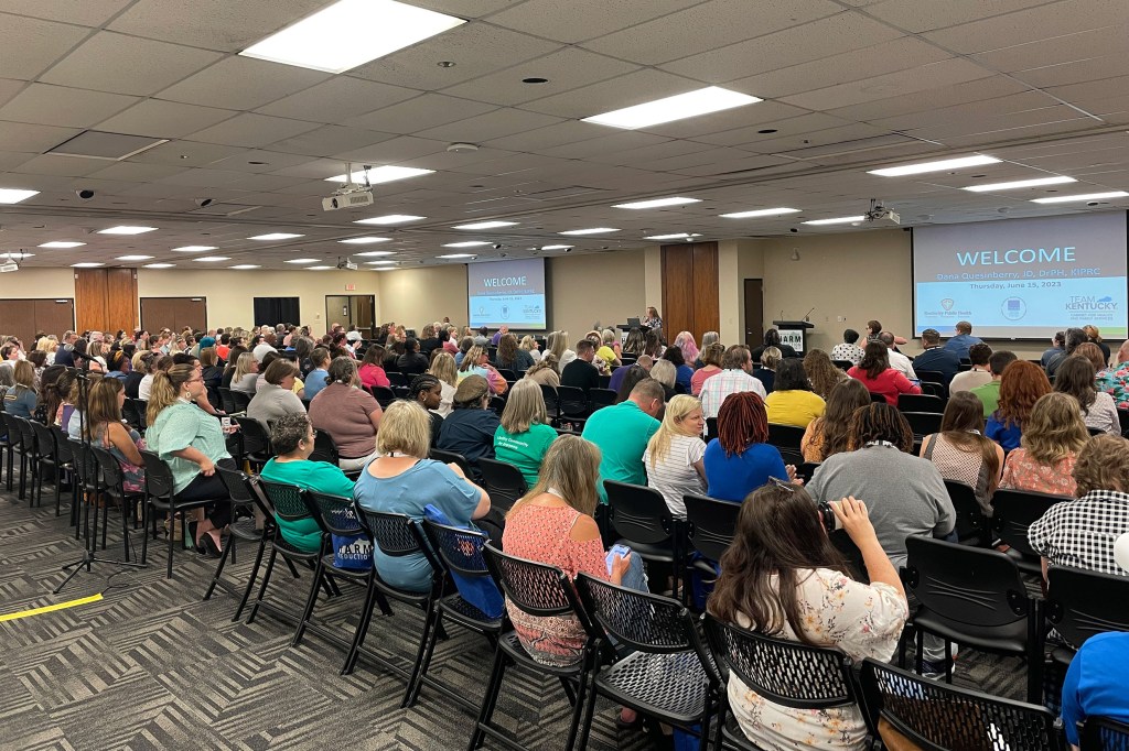 Vital Strategies is hosting a seminar on harm reduction. A large group is seated in rows in a conference room, facing a speaker and presentation screens at the front. The screens display a welcome message. The room is well-lit, with a carpeted floor and dropped ceiling.