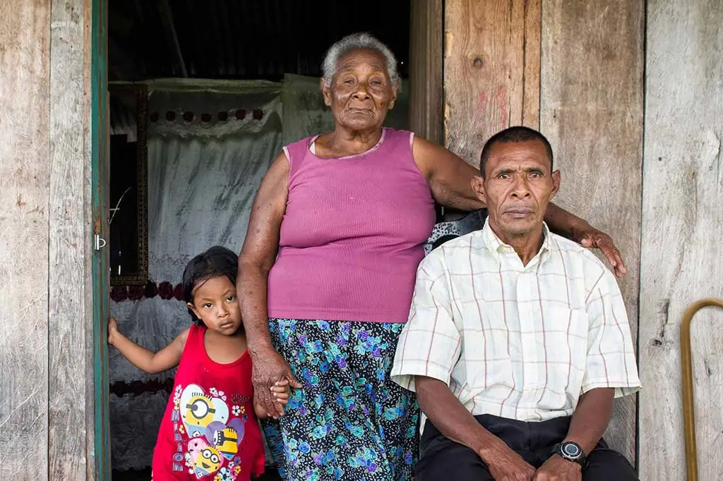 A Honduran man sits in front of a wooden house surrounded by an older woman and a child. There is a cane leaning against the wall behind the man. Photo: Antonio Busiello