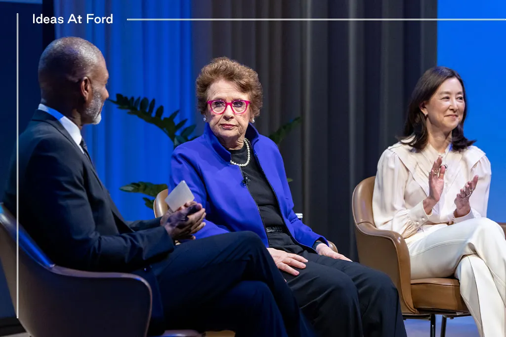 Three people are sitting in conversation against a blue curtain background. The group includes, from left to right, Charles Blow in a dark suit, Billie Jean King in black with a blue blazer, and Clara Wu Tsai in a beige white and trousers.