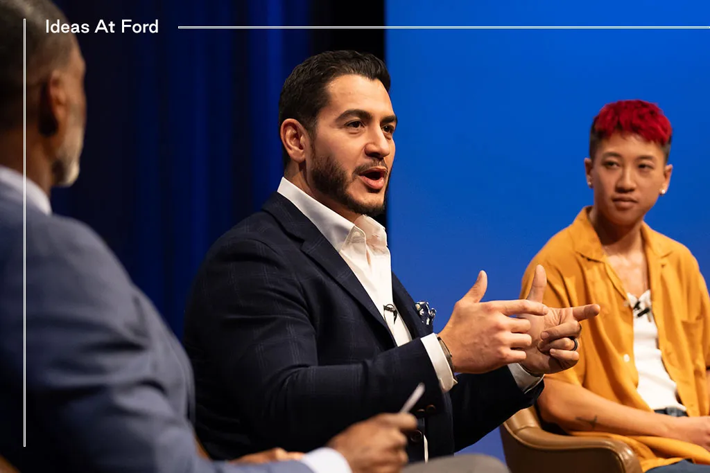 Three people are sitting in conversation against a blue curtain background. From left to right, they are Charles Blow in a blue suit, Dr. Abdul El-Sayed, who has a beard and wears a dark suit with a white collar shirt, and Jes Tom, who has short hair and wears a brown shirt.