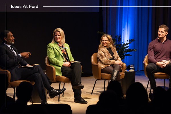 A panel of four people sit on stage during the Ideas At Ford event. Three are smiling as one speaks. They are seated in chairs with a blue-lit backdrop behind them. Left to right they are Charles Blow, Liz Shuler, Jessica Berman, and Carl Nassib.