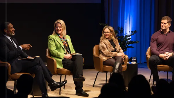 A panel of four people sit on stage during the Ideas At Ford event. Three are smiling as one speaks. They are seated in chairs with a blue-lit backdrop behind them. Left to right they are Charles Blow, Liz Shuler, Jessica Berman, and Carl Nassib.