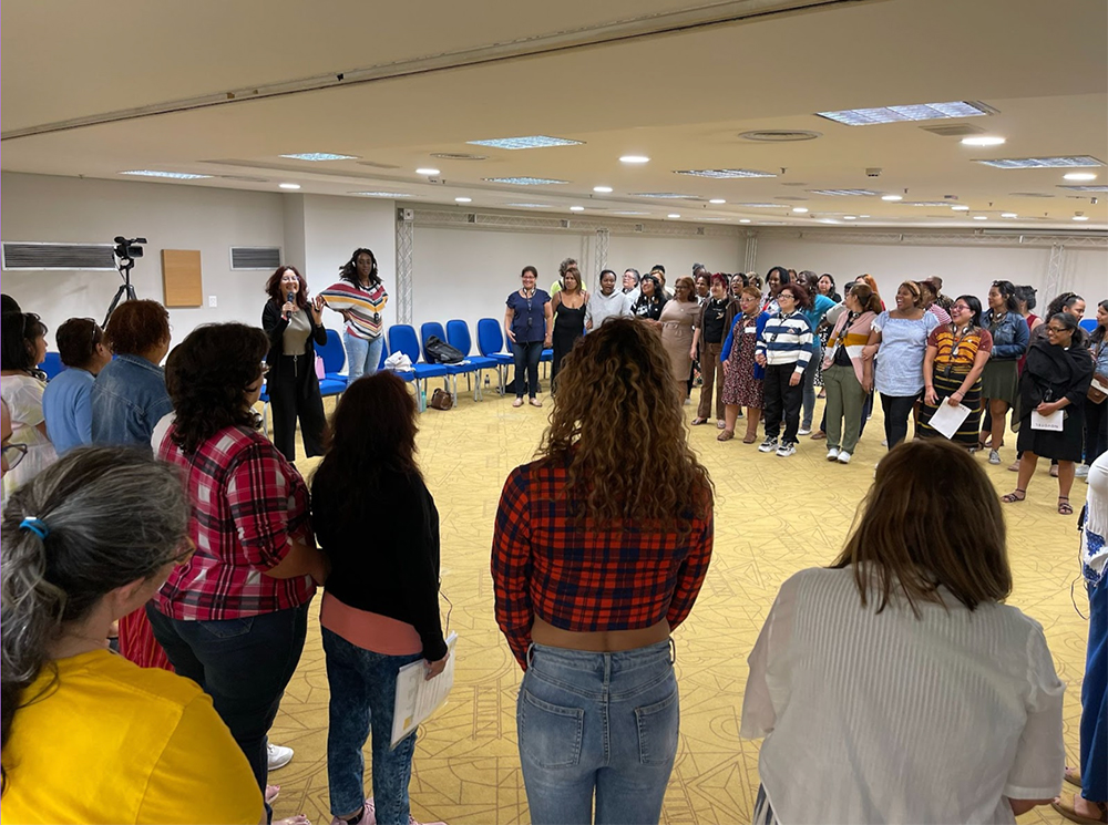 In a well-lit room, participants stand in a circle during the 2023 São Paulo, Brazil LUNA reunion. Some women hold papers, and one woman in the circle speaks in a microphone. Additional chairs and a camera on a tripod are visible in the background.