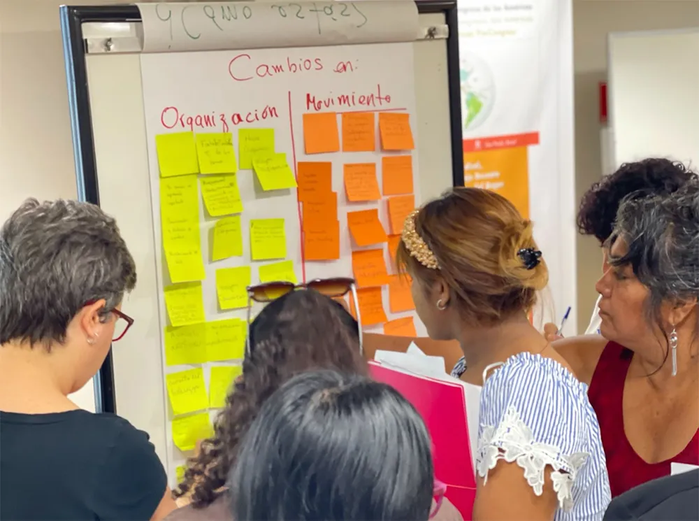 A group of people gather around a flip chart covered with multicolored sticky notes. The notes are under the headings "Cambios en Organización" and "Movimiento." They are discussing changes in labor organizations versus movement ideas in what appears to be a collaborative workshop or meeting.