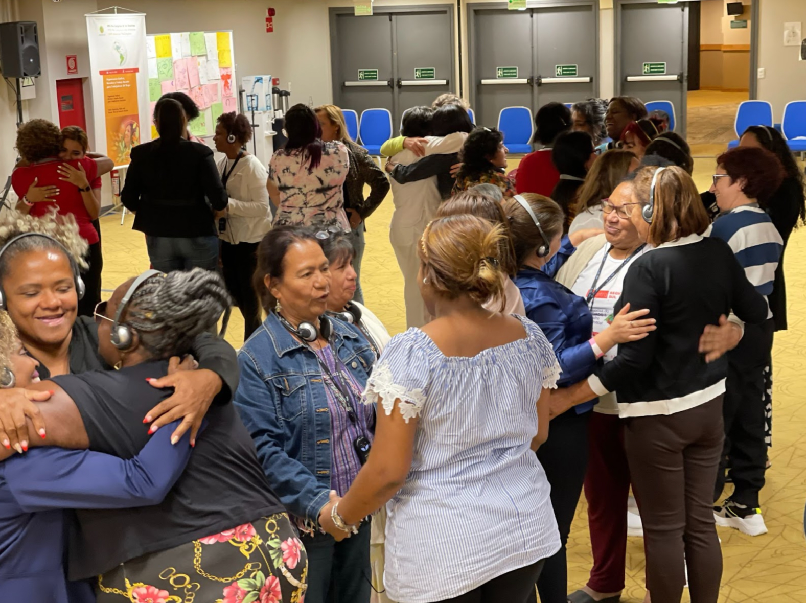 Leaders and affiliates of labor movements worldwide, some with headphones, are standing and hugging each other in a well-lit room with blue chairs in the background. They appear to be conversing and embracing during the 2023 Brazil LUNA reunion, exhibiting warm smiles and camaraderie.