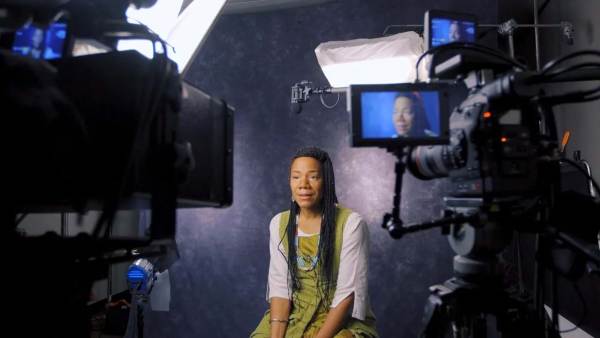 Martha Redbone sitting on a film set in front of a camera with a bright light shining on her.