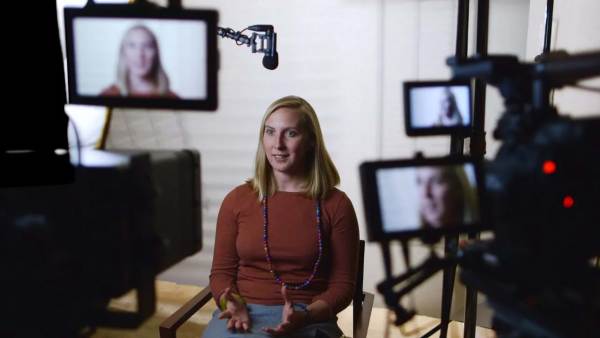 May Boeve sitting on a film set in front of multiple cameras with her face displayed on all monitors.