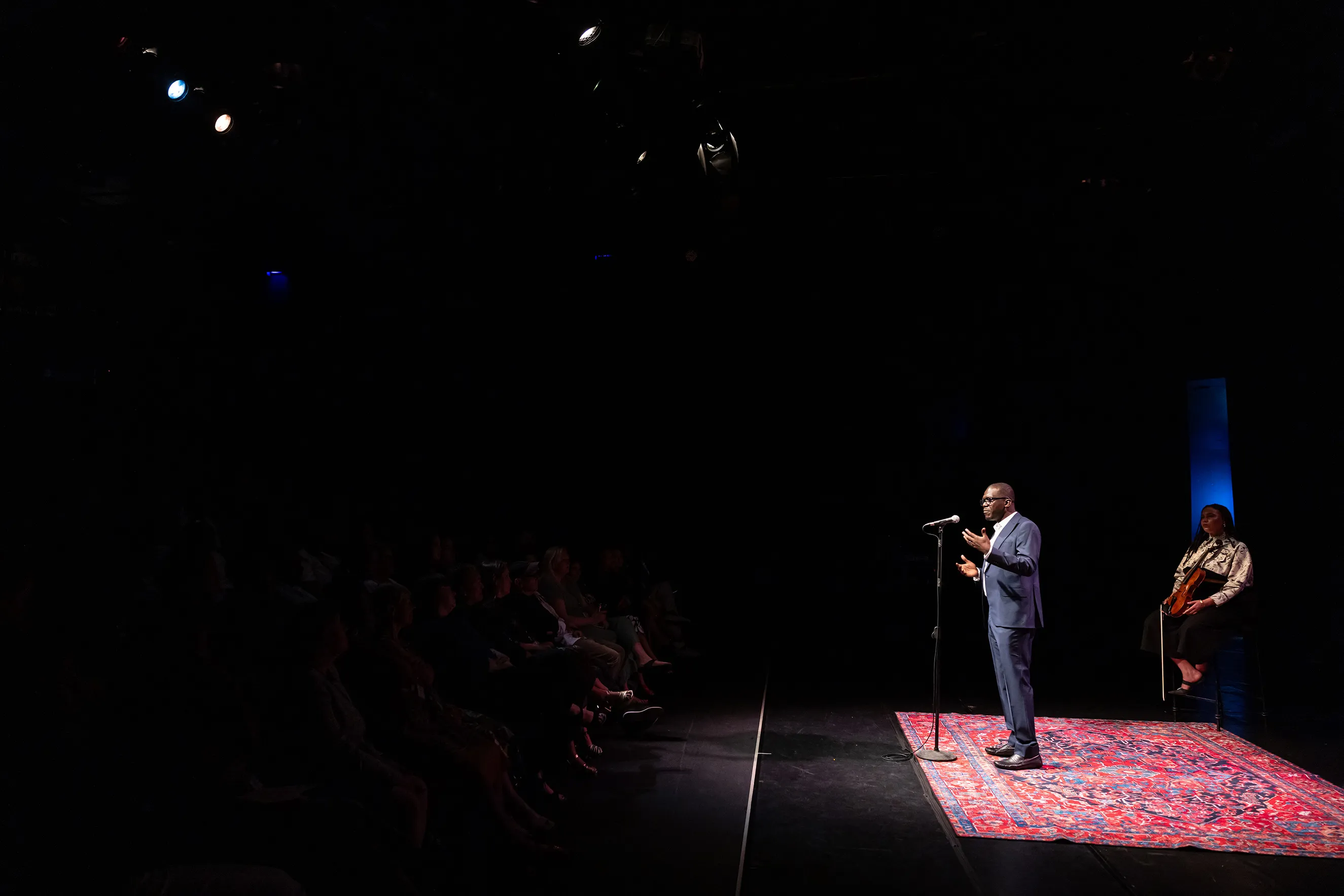 Performer at the microphone wearing a blue suit at the Moth storytelling event at the Ford mainstage at The Ledesi at Joburg Theatre. A violist, wearing black and white, is sitting on a stool in the background.