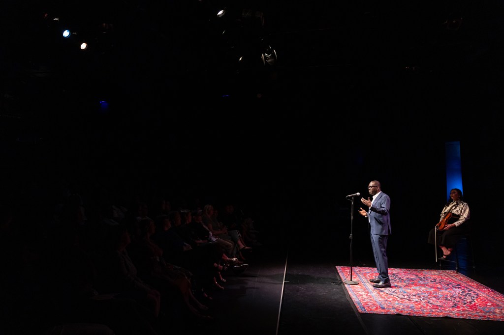 Performer at the microphone wearing a blue suit at the Moth storytelling event at the Ford mainstage at The Ledesi at Joburg Theatre. A violist, wearing black and white, is sitting on a stool in the background.