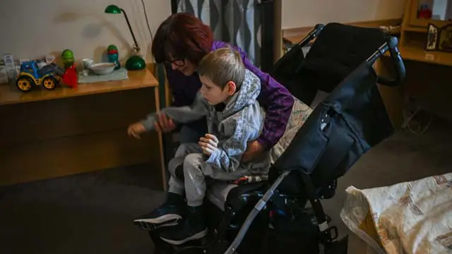 A woman wearing a purple sweatshirt, moves her grandson who suffers from Neurological paralysis from a wheel chair at the Green Hotel in Jerzmanowice, Poland.