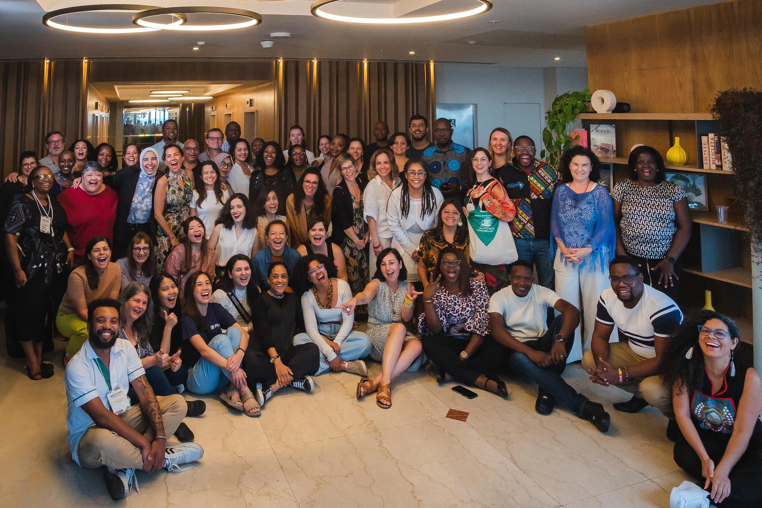 A large, diverse group of people pose together indoors, smiling and laughing. Some are seated on the floor, others standing behind, in a warm, well-lit room with modern décor and bookshelves in the background.