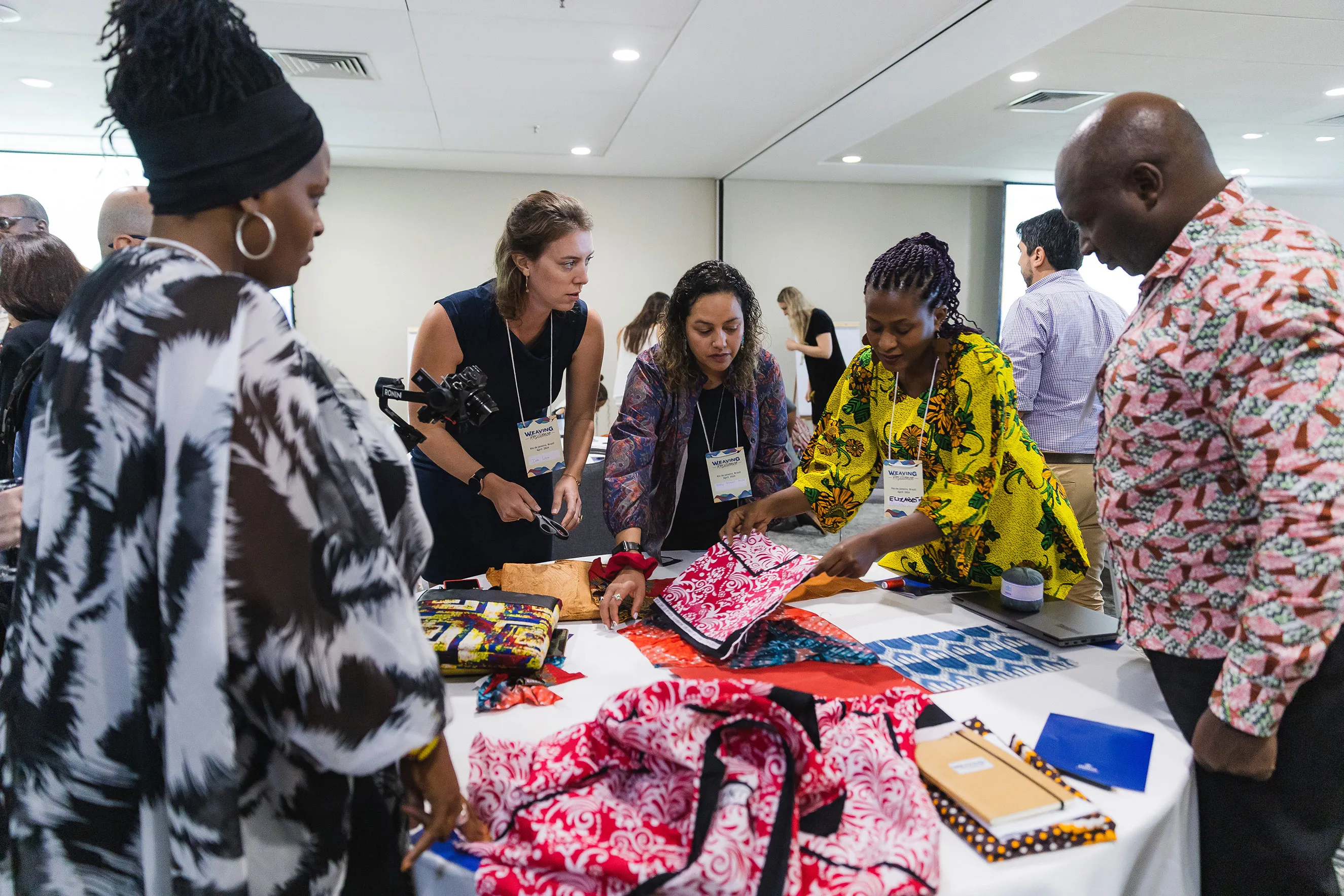 A group of people at a weaving resilience convening gathered around a table display, examining colorful patterned fabrics and handmade items at a conference or workshop in a bright, modern room.