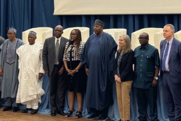 A group of eight people, including men and women in formal and traditional attire, stand side by side indoors in front of a blue curtain and white chairs, posing for a group photo.